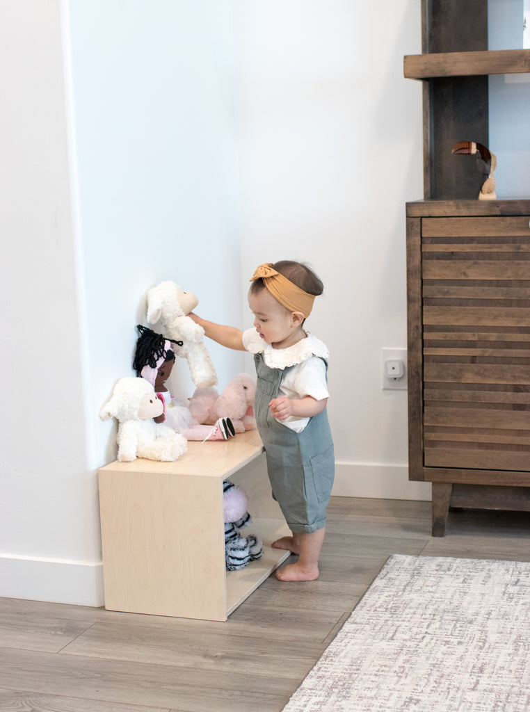 Toddler sets stuffed animals down on her infant montessori shelf (short).