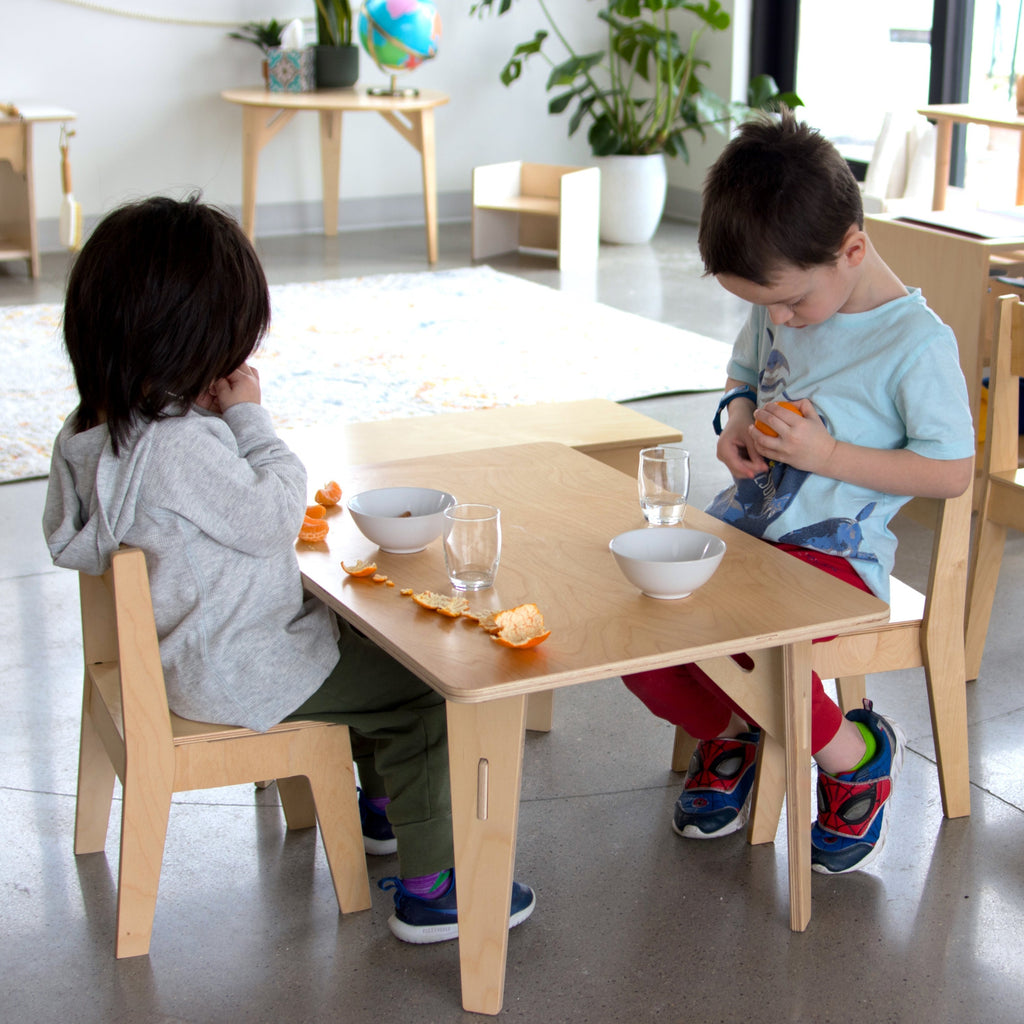 Two young boys eat oranges and drink water sitting at a 20x20 12in high rectangular table.