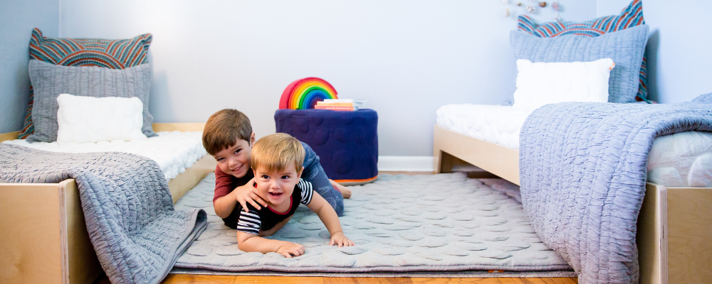 Two children playing on a bed in a child-friendly bedroom with colorful pillows and a rainbow toy.