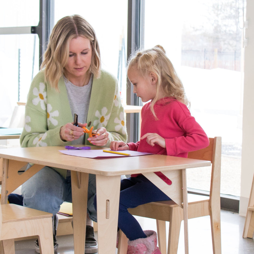 Guide helps young girl learn how to use scissors at a 20x20 Wood Grain Laminate rectangular table.