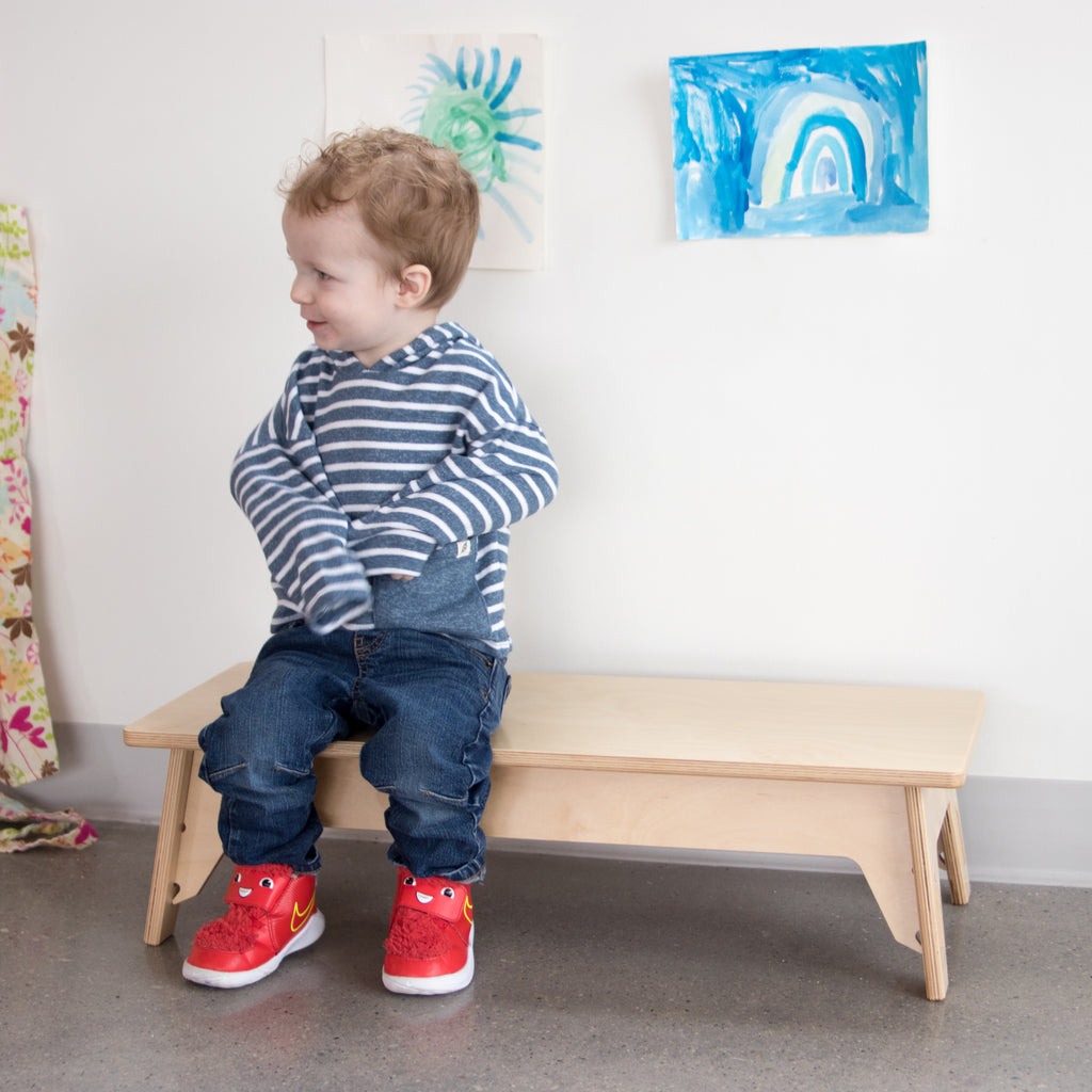 Child in blue top, blue jeans, and red shoes sitting on a low school bench. Artwork from children are posted on the wall behind.