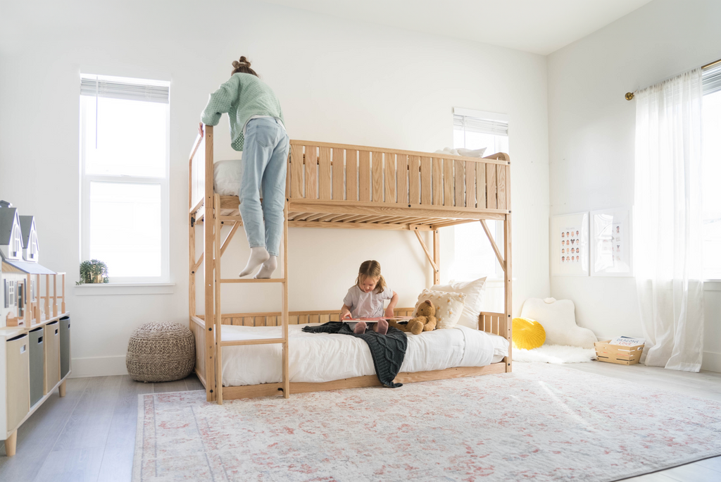 One girl climbs the ladder of the Kids Bunk bed while a younger girl sits on the floor bed bunk bed and reads a book