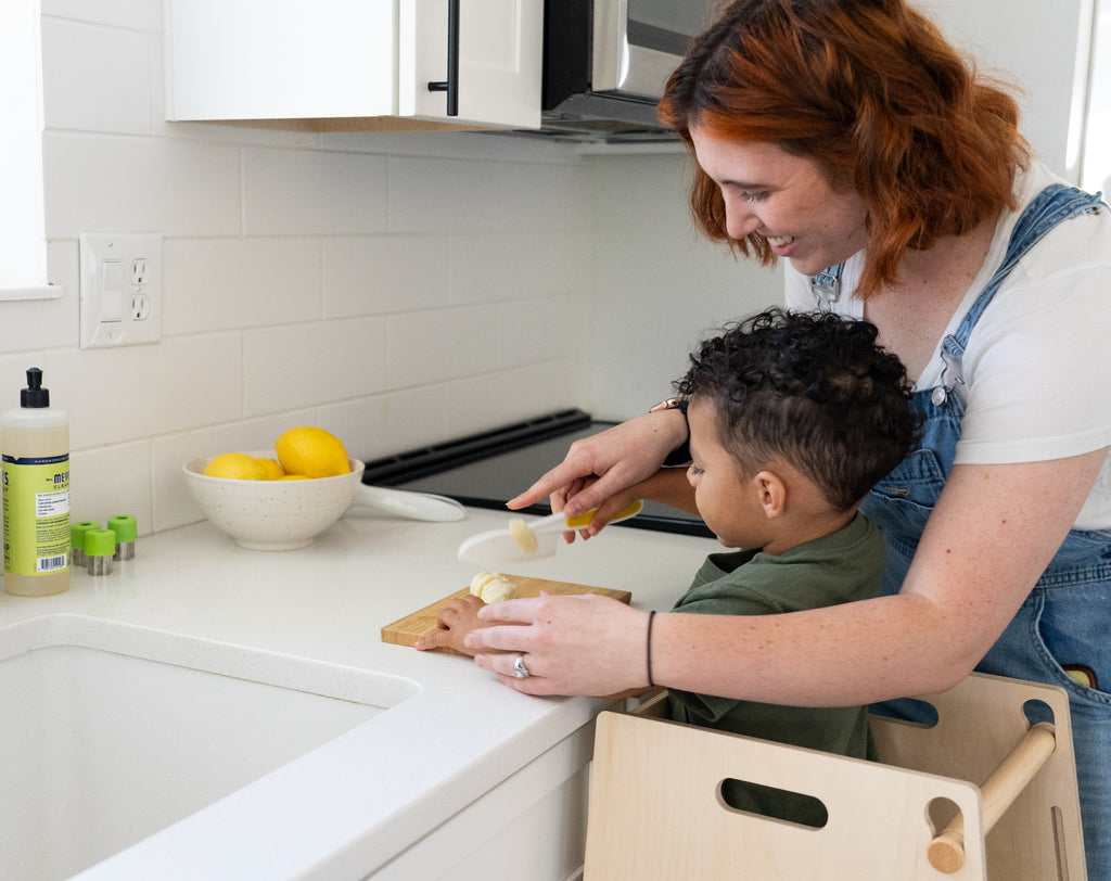 toddler standing at counter in a toddler tower with mom using toddler knife to cut a banana