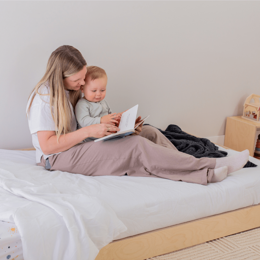 Mom and baby sitting on a floor bed reading a book together.