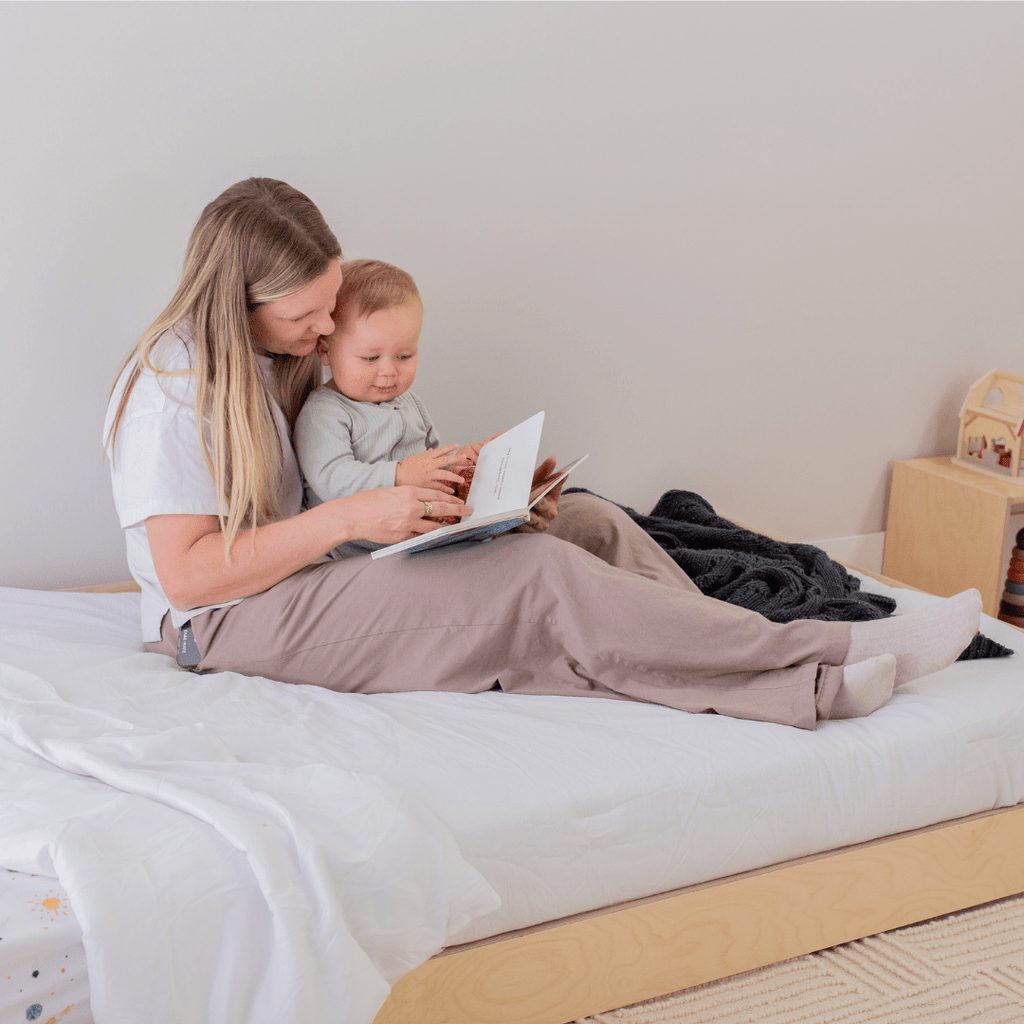 Mom and baby sitting on a floor bed reading a book together.
