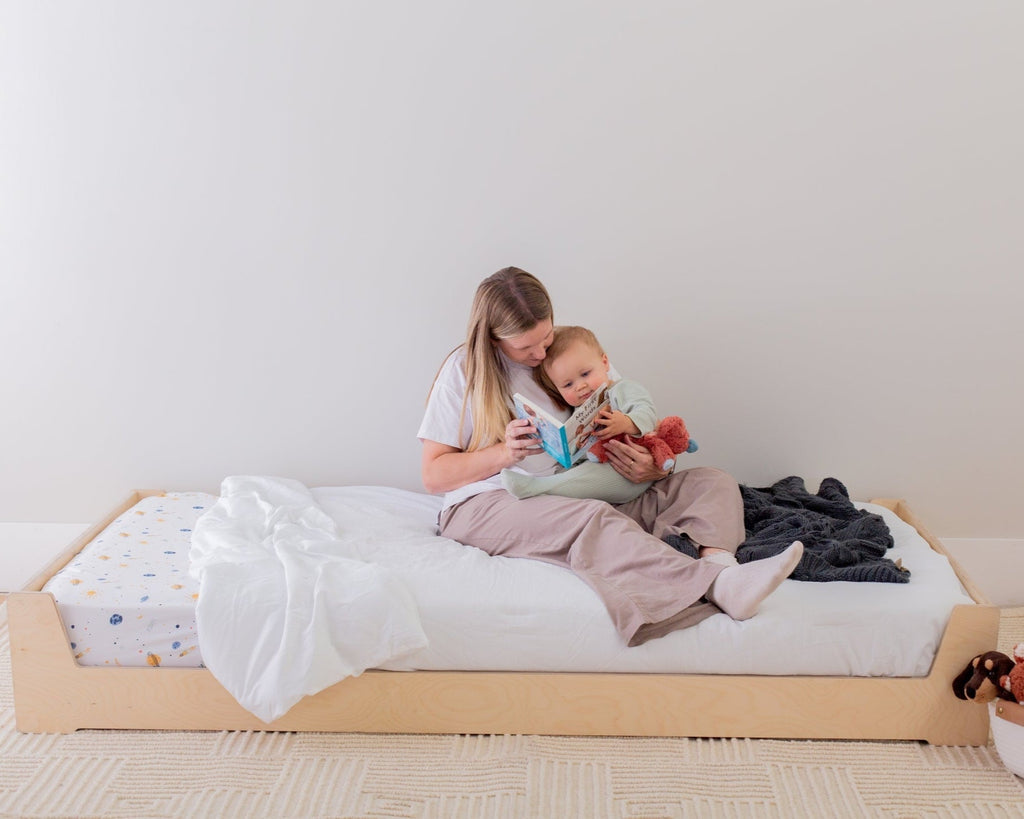 Woman and child sitting on a bed with a white mattress, surrounded by toys and clothes.