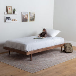 Simple Wooden bed frame with white bedding in a room with a rug and wall art.