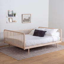 Wooden bed with white bedding and a dark pillow in a room with a white wall and wooden floor.