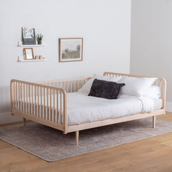 Wooden bed with white bedding and a dark pillow in a room with a white wall and wooden floor.