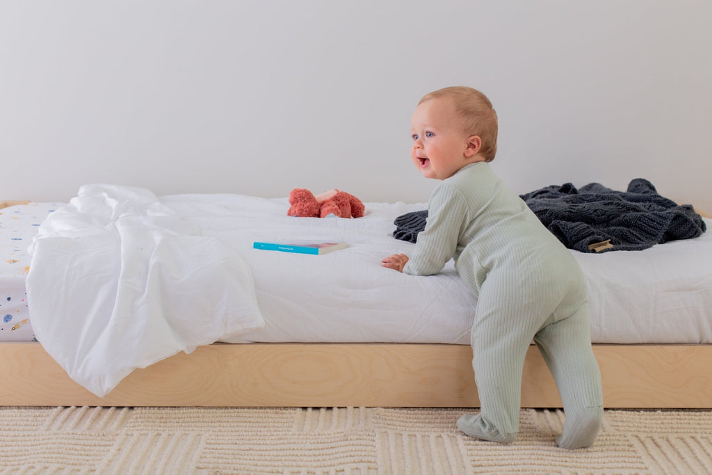 Baby standing by a floor bed with clothes scattered around, in a room with a plain white wall.