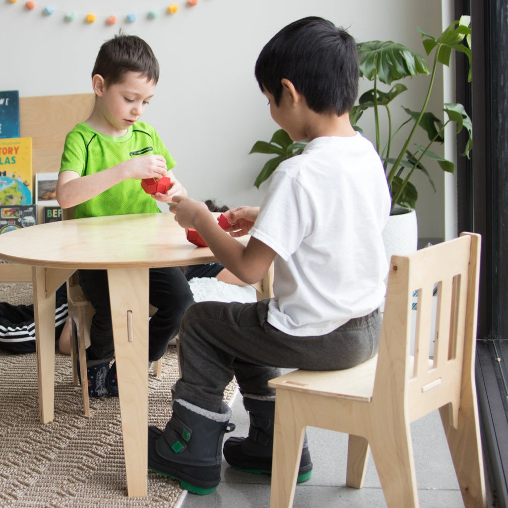 Two children learning at a wooden round table sitting on school kids chairs.