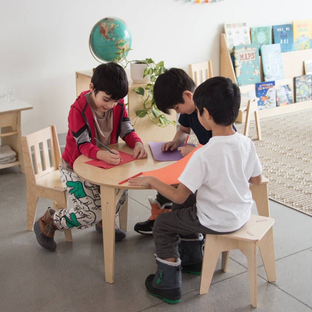 Three young boys sit at a round wooden table on school kids stools and a school kids chair.
