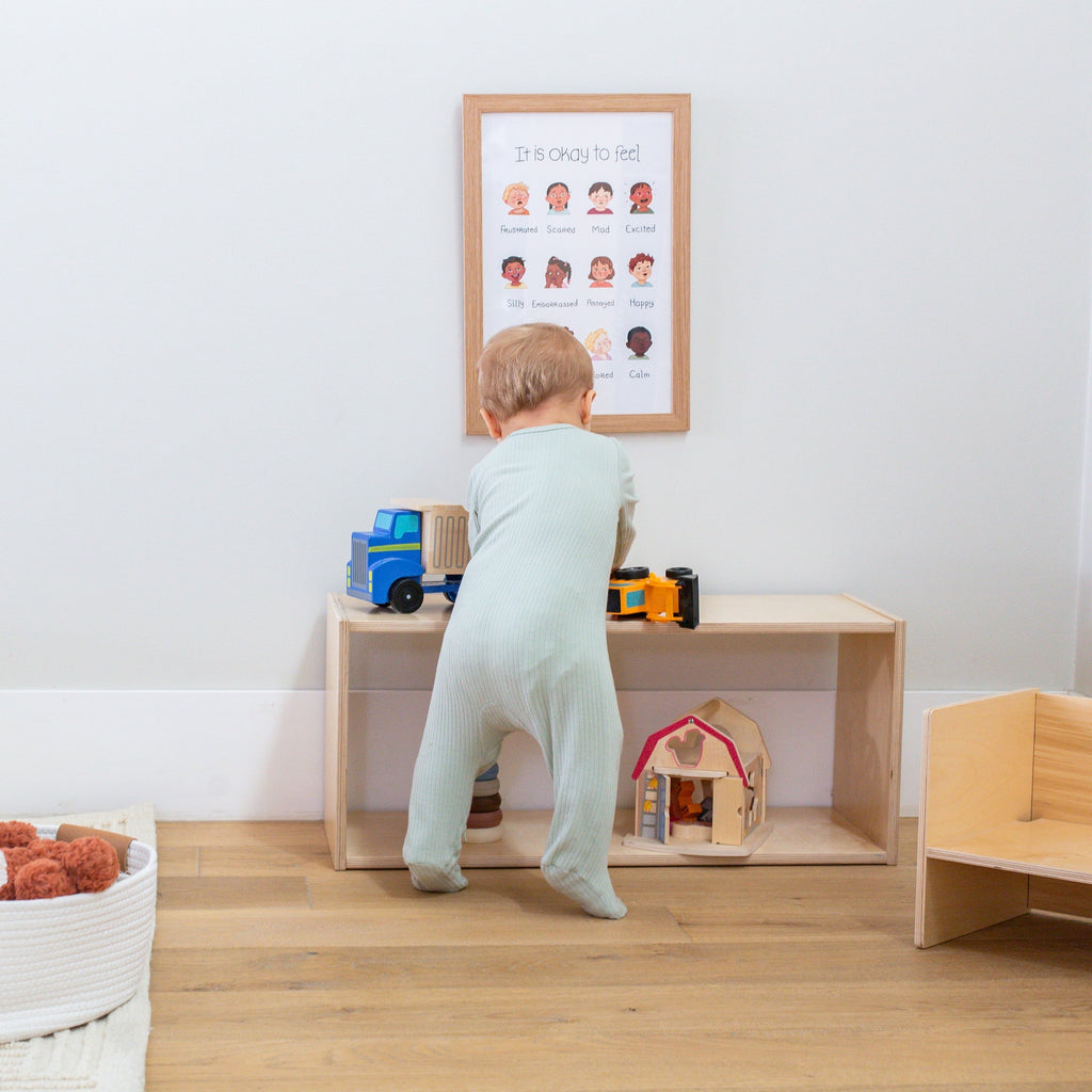 Child playing with toys on a wooden shelf in a room with a framed poster on the wall.