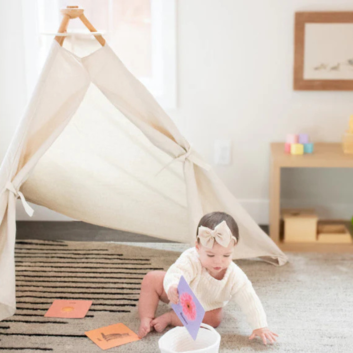 Baby playing with cards near a teepee in a room with a shelf and framed picture.