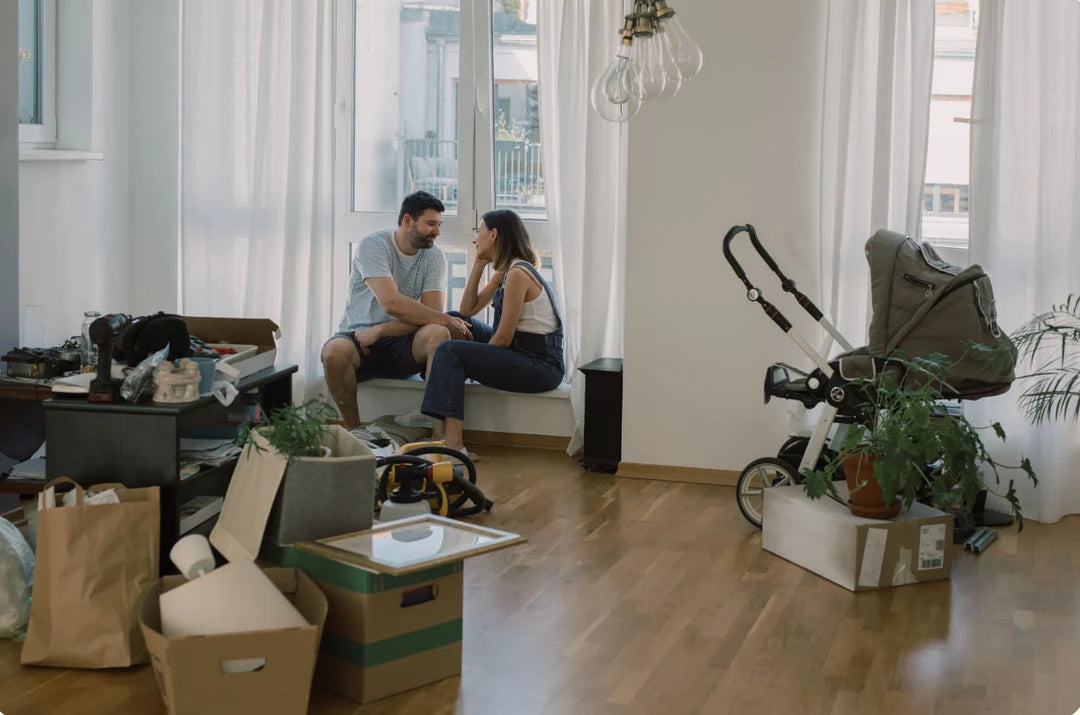 Two people sitting on a couch in a living room with boxes, furniture, and a baby stroller.