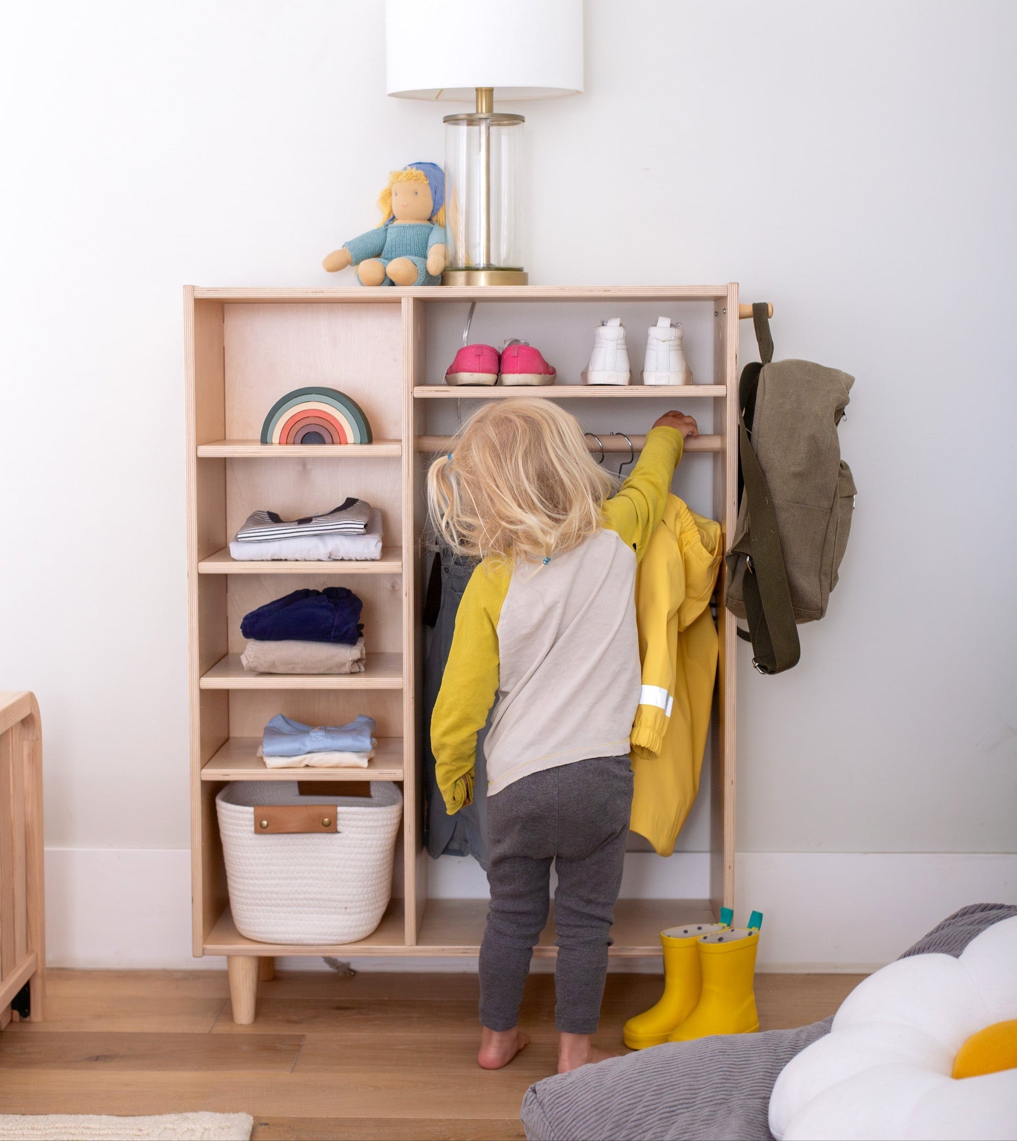 Child standing in a room with a wooden wardrobe and various clothing items displayed and hung up.