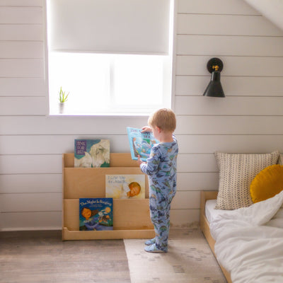 Child in pajamas reading a book in a bedroom with a bookshelf and bed.