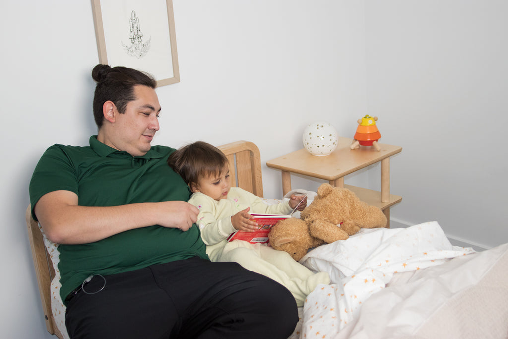 A father and child sit on the toddler floor bed with one headboard as they read a book
