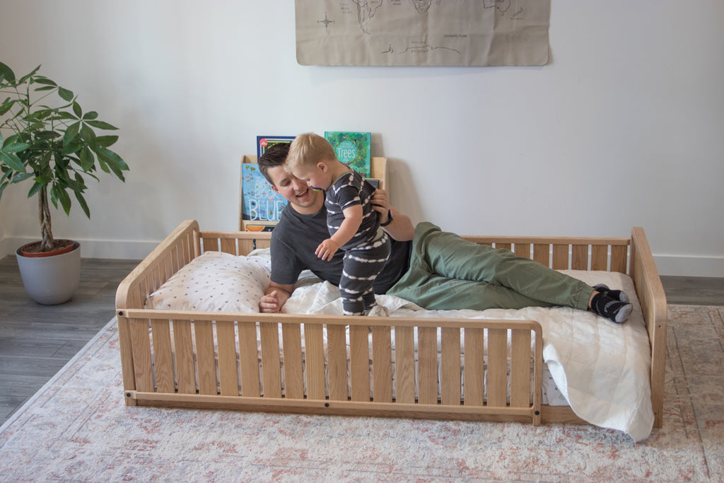 A father and son lay on the toddler floor bed with two headboards, a full side rail, and a 3/4 side rail