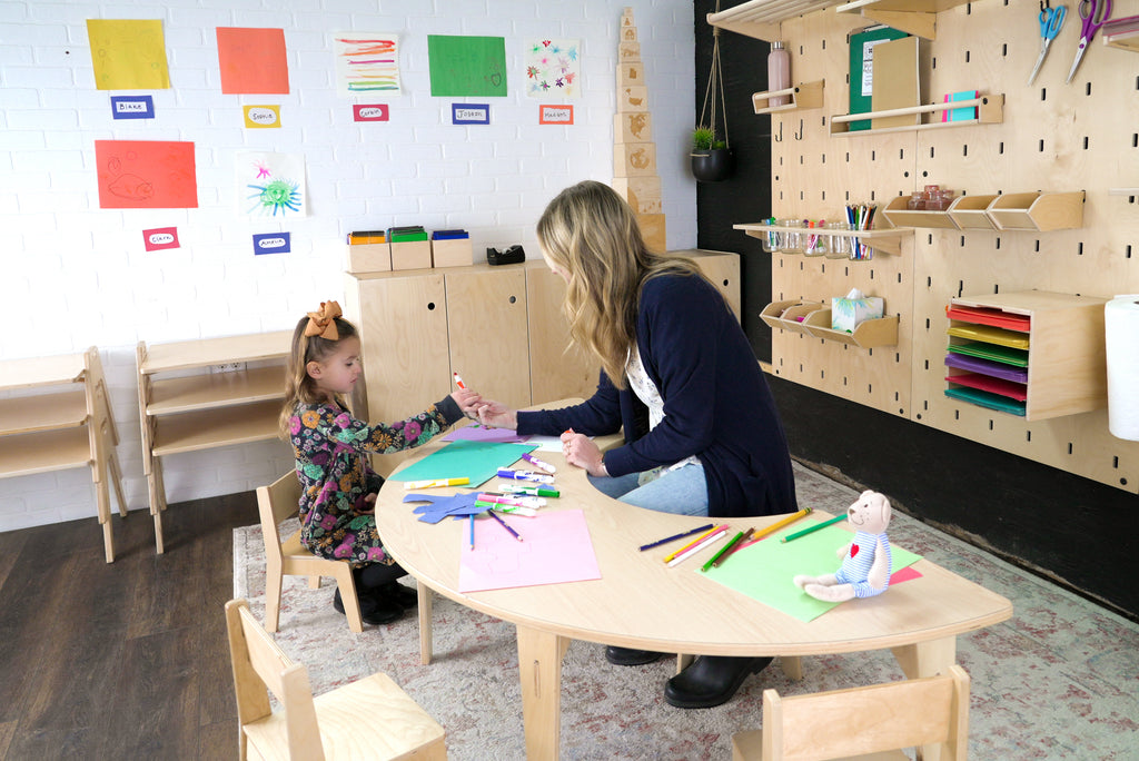 Montessori guide assisting a young girl while she draws at a half round table in a prepared learning environment.